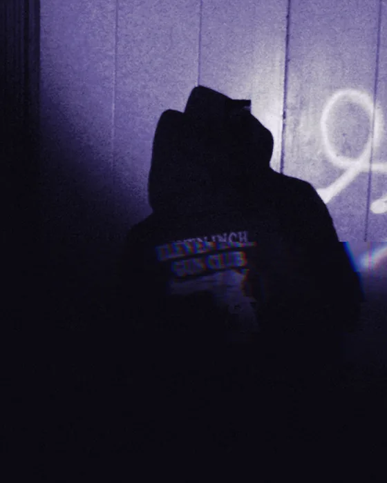 a hooded figure from behind in a flashlight spot on a tagged wall, eleven inch gun club print on the back
